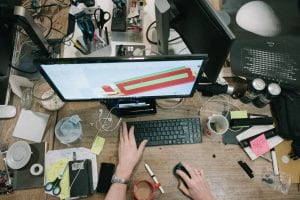 Person working on a computer at a very cluttered desk; image by Robert Bye, via unsplash.com.