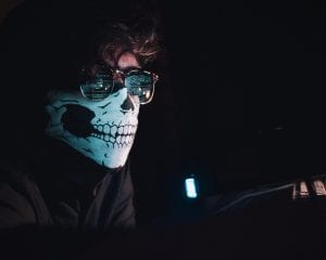 Man in a dark room wearing a skull half-mask and glasses, data from a computer screen reflected in his glasses; image by Nahel Abdul Hadi, via Unsplash.com.