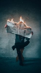 Man sitting on stool reading a newspaper that is on fire; image by Elijah O&rsquo;Donnell, via Unsplash.com.