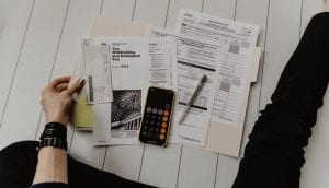 Woman in black pants sitting on floor with tax paperwork and a calculator; image by Kelly Sikkema, via Unsplash.com.