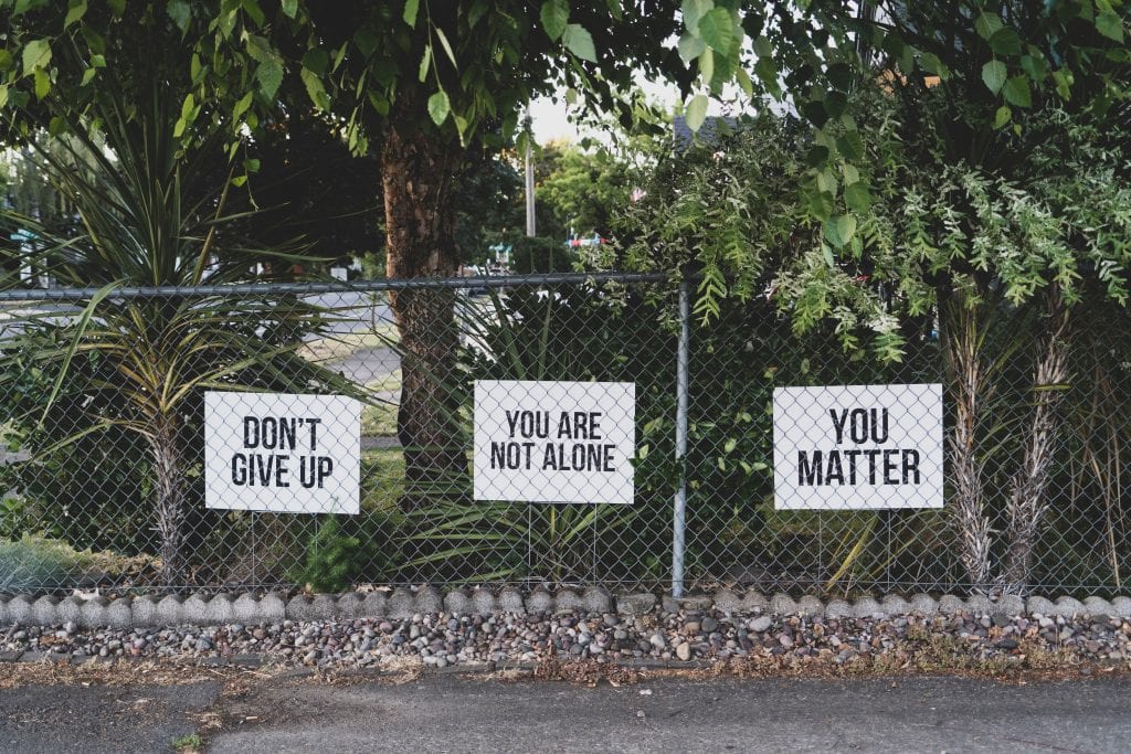 Signs by fence line: &ldquo;Don&rsquo;t give up,&rdquo; &ldquo;You are not alone,&rdquo; &ldquo;You matter.&rdquo; Image by Dan Meyers, via Unsplash.com.