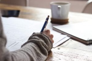 Person in grey sweater with notebook, pen, and papers, with mug in background; image by Green Chameleon, via Unsplash.com.