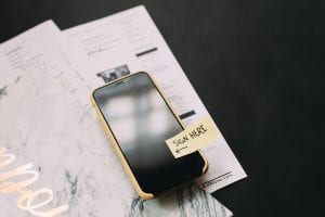 Black smartphone on top of papers. Phone has a sticker on the screen saying &ldquo;Sign here.&rdquo; Image by Kelly Sikkema, via Unsplash.com.