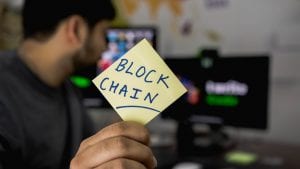 Man holding sticky note with &ldquo;Blockchain&rdquo; written on it; image by Hitesh Choudhary, via Unsplash.com.