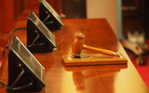 A gavel lays on a wooden table with monitor screens nearby.