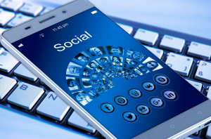 Smartphone with &ldquo;Social&rdquo; on the screen & various social media icons, sitting on computer keyboard; image by Geralt, via Unsplash.com.