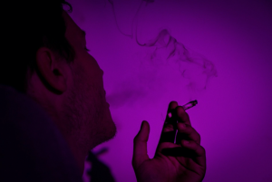 Profile view of man smoking in purple light; image by GRAS GR&Uuml;N, via Unsplash.com.