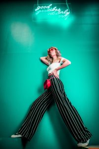 Woman in striped pants against green background with &ldquo;Good Vibes Only&rdquo; neon sign; image by Ahmed Carter, via Unsplash.com.