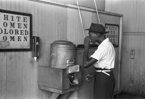 A black man sips from a cup at a segregated drinking area.