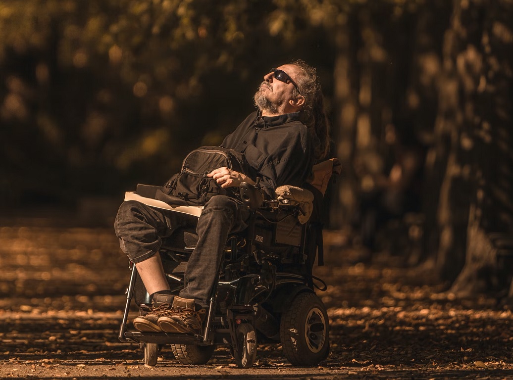 A man wearing sunglasses, seated in a mechanical wheelchair in a natural area.